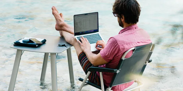 A man built his camping table and chair in a river and works on his laptop.