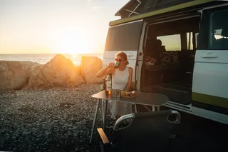 A woman sits on a shore in front of an open camperBoys camper at the camping table and drinks from a cup.