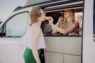 A woman standing inside a camper behind the built-up kitchenette and lets another woman standing outside bite off a muesli bar.