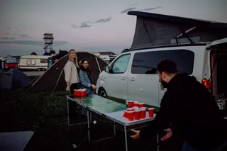 Two women play a round of beer pong against a man, in the background you see a festival ground with tents and a Opel Crosscamp. You can read what you need for the Festival Camping at our Festival Packliste.